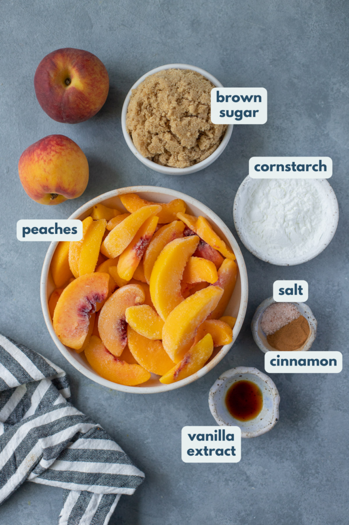 Labeled bowls of ingredients on a gray background: frozen peaches, vanilla extract, salt, cinnamon, brown sugar, and cornstarch.