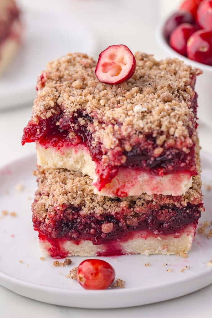 Two homemade cranberry bars stacked on a white dessert plate. The bar on top has a bite taken out of it.